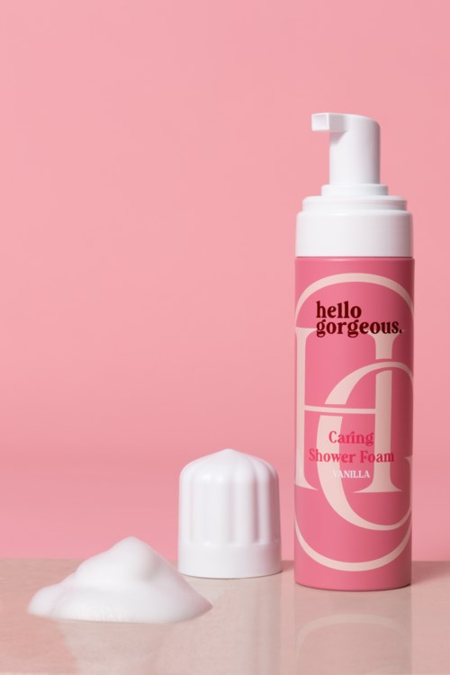 En rosa flaska med duschskum, vit lock och lite skum bredvid, på rosa och grå bakgrund.