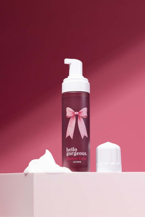 A pink bottle of shower foam with a bow label and foam beside it on a pink background.