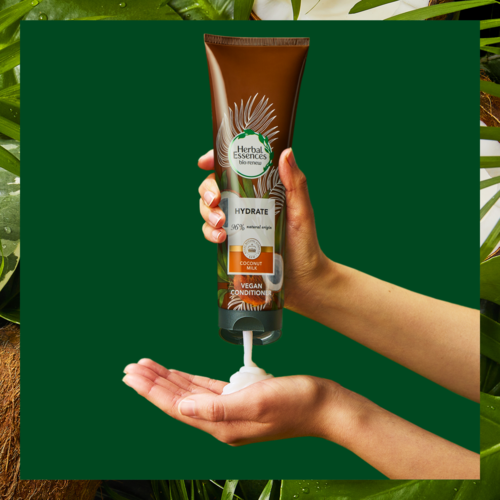 En hånd holder en brun flaske merket 'Herbal Essences', med kokosmelk, vegansk balsam, og en hvit krem som blir utløst.