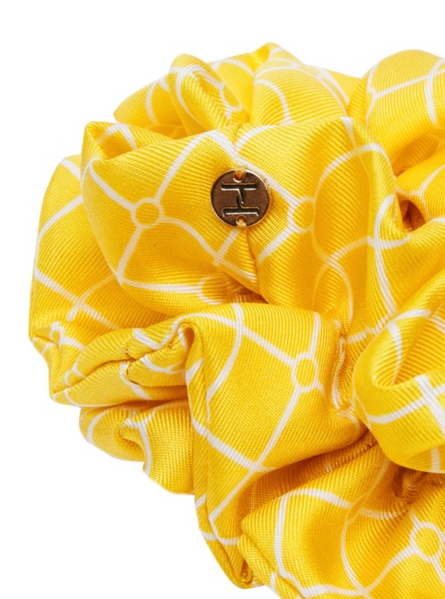 En gul tygscrunchie med en liten metalllogo och vit geometrisk mönster. Den är tillverkad av mjuk, stretchig material.