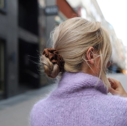 En kvinna med blont hår i en knut, bär en lila tröja, är utomhus på en stadsgata.