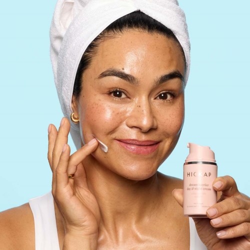 Eine Frau mit Handtuch im Haar trägt Hautcreme auf das Gesicht auf, lächelt. Sie hält eine rosa Flasche.