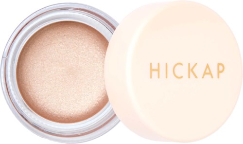 Ein kleiner runder Behälter mit einem beigen, schimmernden Make-up-Produkt und einem weißen Deckel mit goldenen Buchstaben, die "HICKAP" sagen.