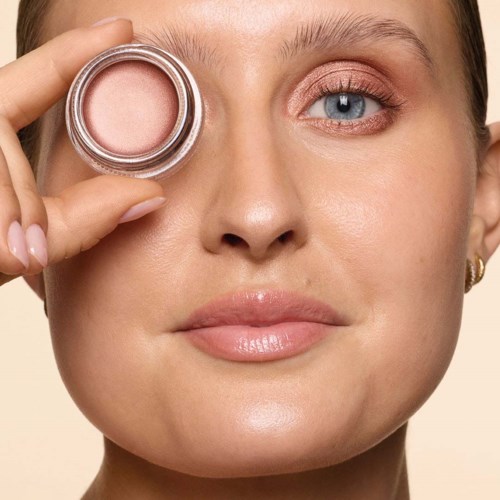 Eine Frau hält ein kleines Glas mit rosa Blush in der Nähe ihres Auges, lächelt leicht mit klarer, strahlender Haut und blauen Augen.