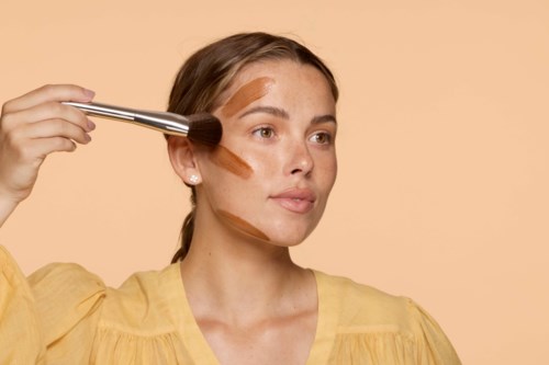 En kvinna applicerar makeup med en stor borste, lite brunt smink syns på hennes ansikte, och hon bär en gul topp.