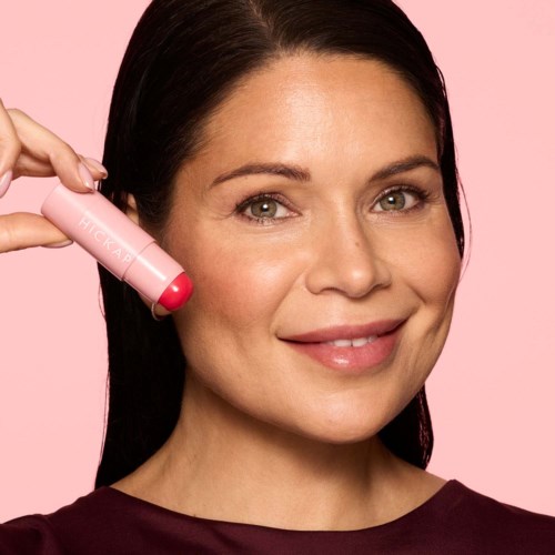 Kvinde smilende, holder en pink kapsel nær ansigtet, mod en pink baggrund.