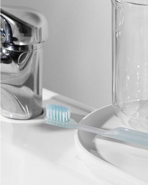 En tannbørste ligger på en tallerken nær et glass, med vask og kran i bakgrunnen. Rent og minimalistisk.