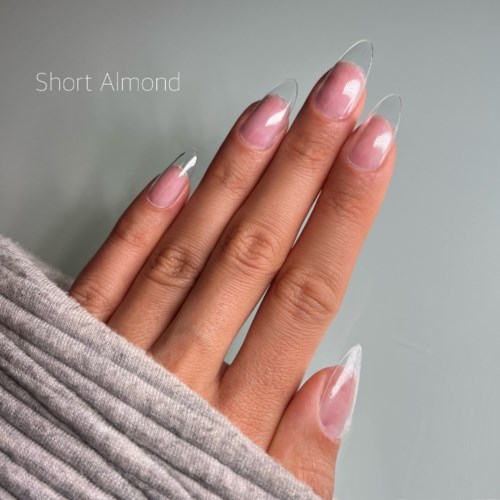 Nude nails with long, clear tips shown on a hand, partially covered by a gray fabric.