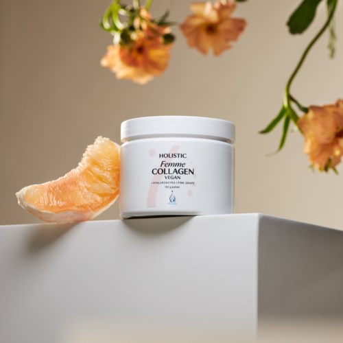 En hvit boks med teksten 'Femme Collagen Vegan' på en hvit overflate, med en rosa frukt og oransje blomster i nærheten.