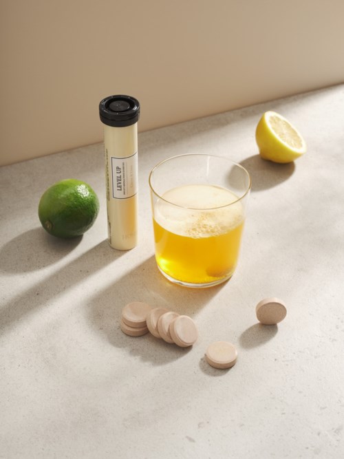 Ett glas öl, en halva citron, en lime, tabletter och en cylinder på en ljus yta.