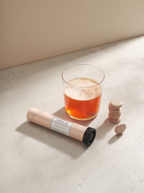 Ett glas med rödaktig dryck, ett ihoprullat papper märkt 'LEVEL UP' och flera vita tabletter på en ljus yta.