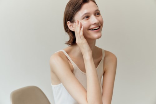 Een lachende vrouw met kort bruin haar zit aan een tafel, raakt haar gezicht met haar hand, in een lichte top.