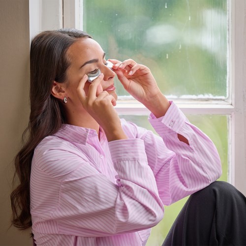A woman in a pink shirt sits by a window, holding a facial roller to her face, relaxing.