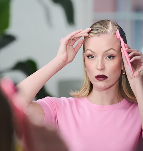A woman with blonde hair, wearing a pink shirt, is using a pink hairbrush to style her hair.