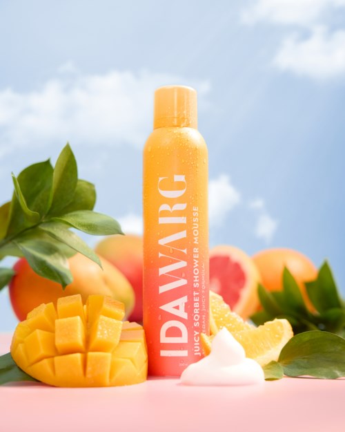En mango-dugskumflaske omgivet af mangoskiver, blade og citrusfrugter under blå himmel.
