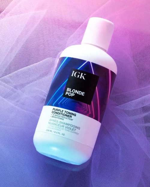 En hvit flaske med IGK Blonde Pop lilla tonende balsam på lilla og rosa stoffbakgrunn.