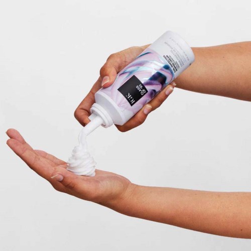 Eine Person gießt Lotion oder Creme aus einer weißen Flasche in ihre Handfläche vor einem schlichten Hintergrund.