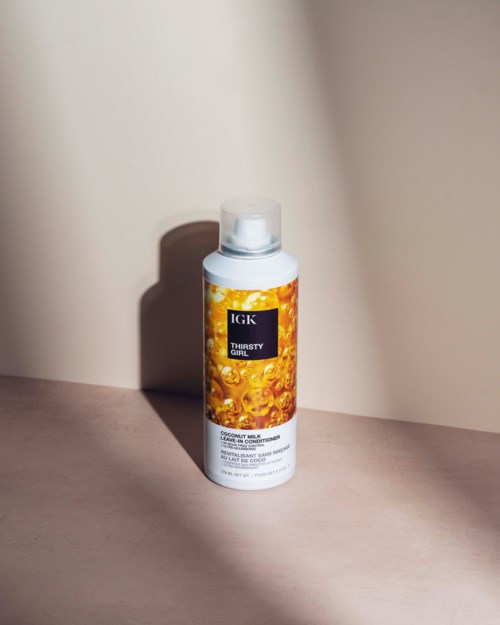 En vit flaska märkt 'IGK Thirsty Girl' med orange vätska står på en rosa yta och kastar skugga.