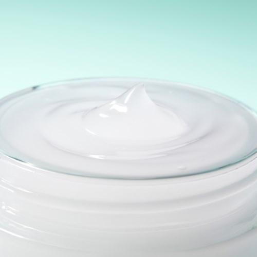 A close-up of white cream or lotion in a glass jar against a light background.
