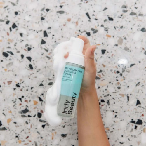 Eine Hand hält eine Flasche mit Schaumreiniger, aus der weißer Schaum austritt, vor einem gepunkteten Hintergrund.