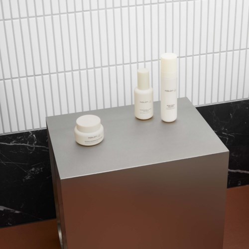 Drie witte cosmetica-flessen en een pot staan op een grijze tentoonstellingsplank tegen een witte betegelde muur.
