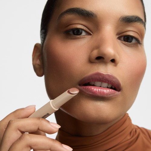 En kvinna som applicerar läppstift, nära bild av hennes läppar och ansikte med neutral makeup, framför en enkel bakgrund.