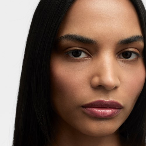 En närbild av en kvinna med rakt mörkt hår och naturlig makeup mot en ljus bakgrund.