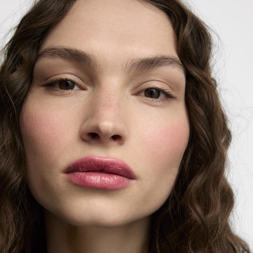 En närbild av en kvinna med lockigt brunt hår, naturlig makeup och rosa läppar, tittar på kameran.