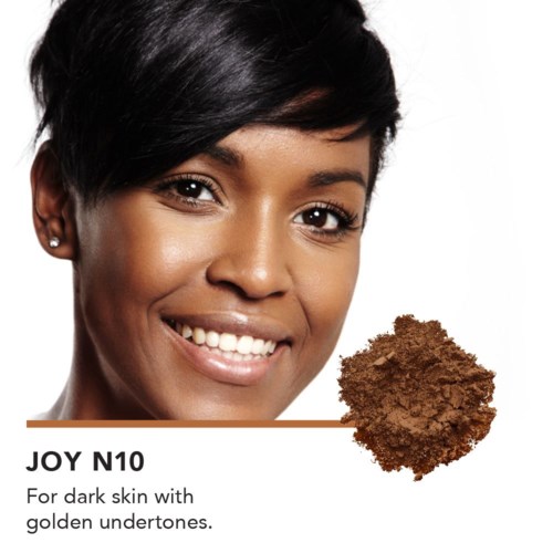 Nainen, jolla on tumma iho ja lyhyt musta tukka, hymyilee. Vieressä on kasa ruskeaa jauhetta. Teksti mainitsee "Joy N10" tummalle iholle kultaisilla sävyillä.