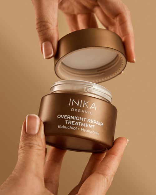 En person åpner en gullfarget krukke merket 'INIKA Organic Overnight Repair Treatment' med synlige hender.