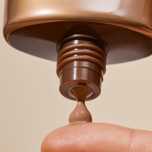En hand med ett finger nära ett brunt rör som lämnar ut en beige kräm.