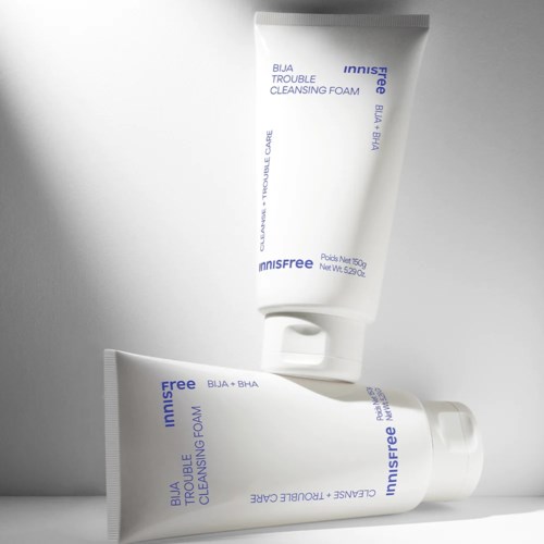Two white tubes of skincare on a gray surface, with blue text indicating they are cleansing foams for trouble skin.