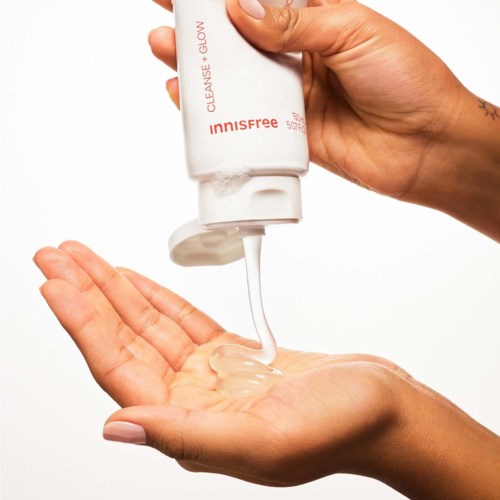 Een hand brengt een wit huidverzorgingsproduct uit een tube op een andere hand, tegen een witte achtergrond.