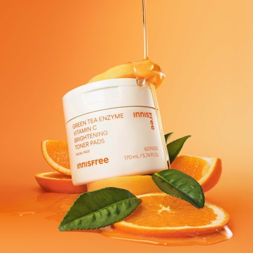 Een pot Innisfree Vitamin C Brightening Toner Pads omringd door sinaasappelschijven en groene bladeren op een warme oranje achtergrond.