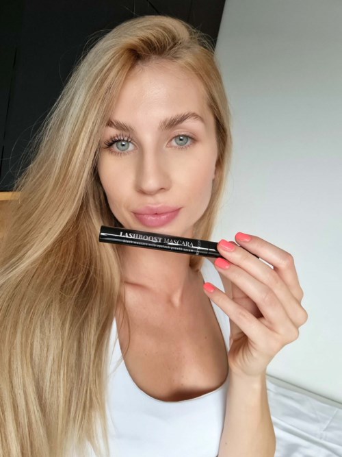 Kobieta z długimi blond włosami trzyma czarną mascarę blisko twarzy, stojąc w jasnym pokoju.