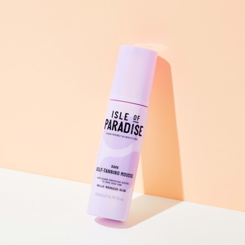 A white bottle labeled 'Isle of Paradise' with purple accents, standing on a pastel background. It is a self-tanning mousse.