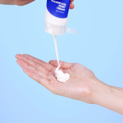 En hånd holder en lille mængde creme fra en flaske mærket 'Hyaluronic Acid Moist Cream' mod en blå baggrund.