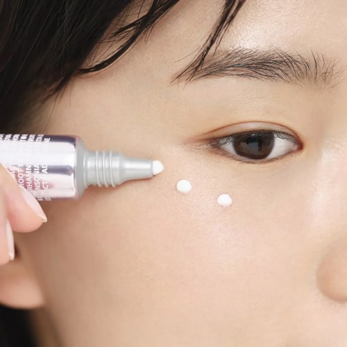Someone applies skincare cream under their eye using a small tube. The person's face is close-up and focused.
