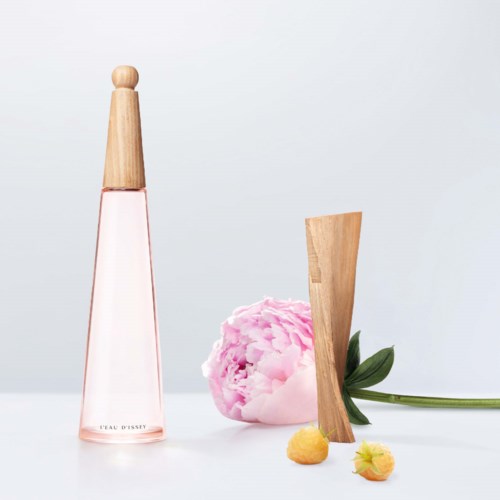 En rosa parfymflaska, en rosa pionblomma, ett träföremål och små gula och rosa frukter på en vit bakgrund.