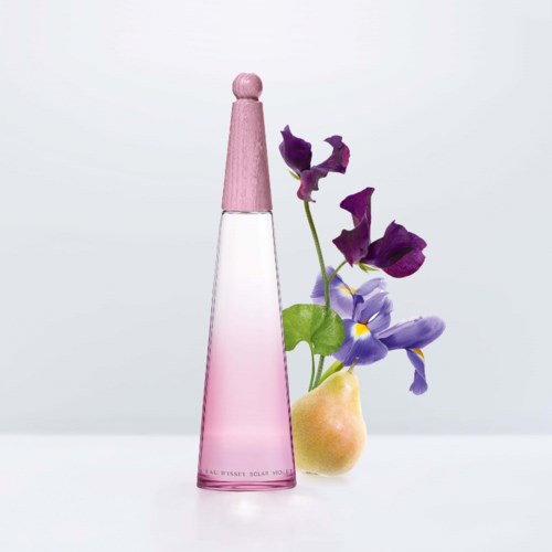 En rosa parfymflaska, gul päron och lila blommor sammansatta på en ljus bakgrund.