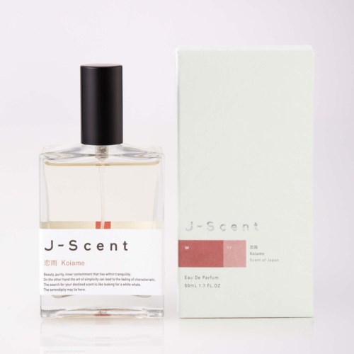 Eine klare Parfümflasche mit schwarzem Deckel und J-scent-Logo neben einem weißen Karton mit minimalistischem Design und hellrosa Details.