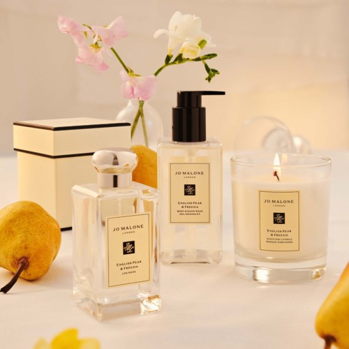 Elegant bathroom setup with perfume bottles, a candle, yellow pears, and pink flowers on a soft background.