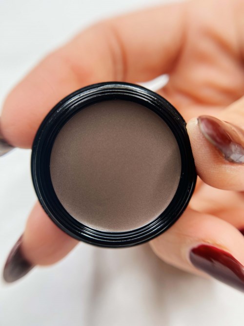 A hand holding a small round container of gray makeup or cosmetic product.