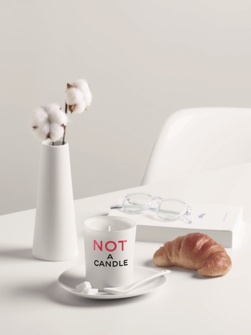 Eenvoudige scène met een witte vaas en katoen, een beker met "NOT A CANDLE," een boek, een bril en een croissant.