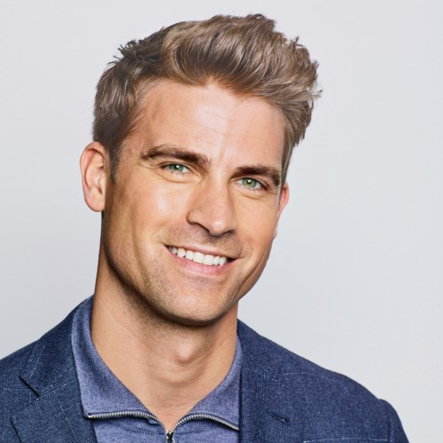 Ein lächelnder Mann mit kurzen, gestylten braunen Haaren, heller Haut, gekleidet in blauen Blazer und Hemd.