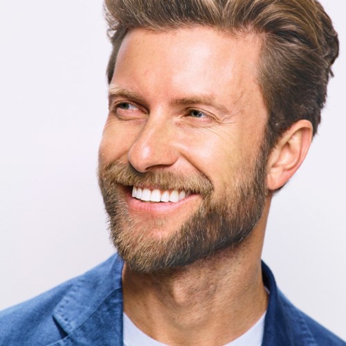 En leende man med skägg och ljusbrunt hår, bär en blå blazer, tittar åt sidan.