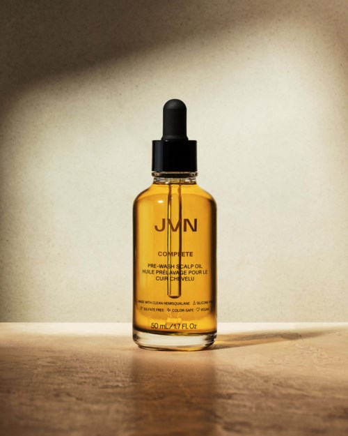 En liten bervaxflaska med pipettlock, märkt 'JMN Complete Pre-Wash Scalp Oil,' står på en träyta.