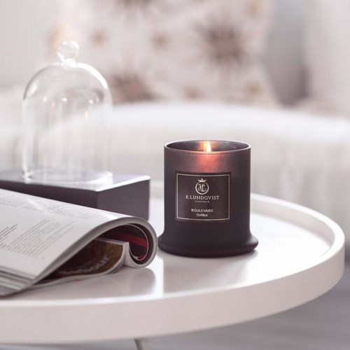 Een brandende zwarte geurkaars met een stijlvolle label op een witte ronde tafel, een open tijdschrift en een glazen stolp op de achtergrond.