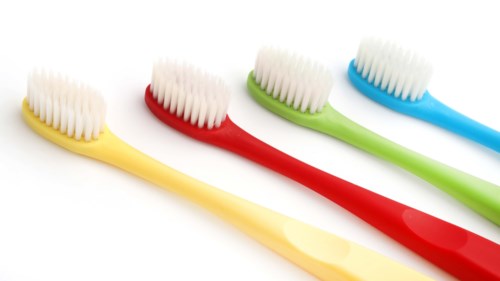 Four colorful toothbrushes are lined up, yellow, red, green, and blue, with white bristles on a white background.