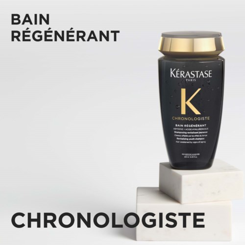 Czarna butelka szamponu z złotą nakrętką z Kerastase Paris stoi na białej kostce. Tekst zawiera "Bain Regenerant" i "Chronologiste."